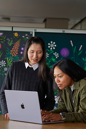 Dos personas del equipo de Marketing de Apple trabajan juntas en una MacBook delante de carteles diseñados por su equipo.