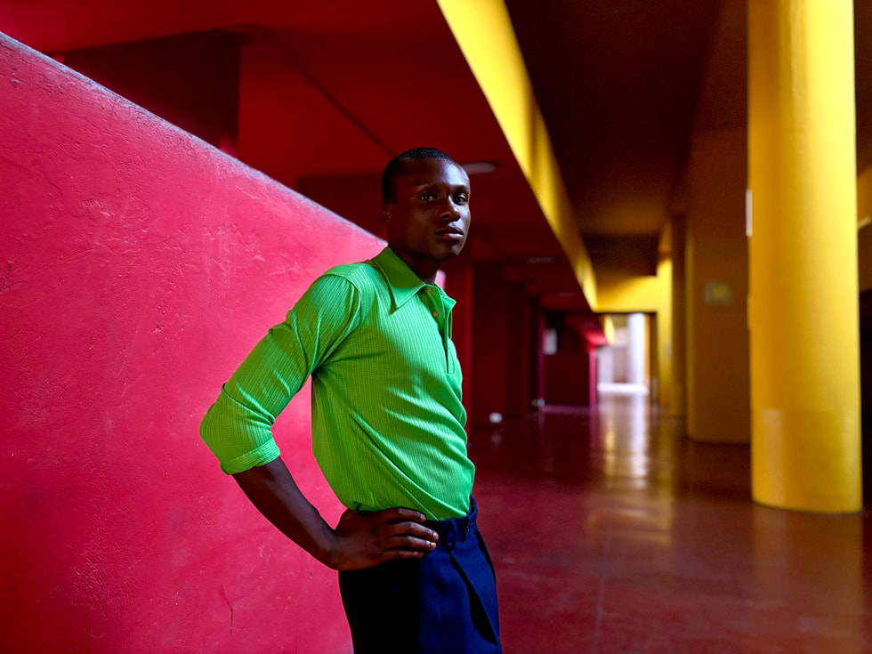 A portrait taken on iPhone 15 shows a person wearing a vibrant green shirt and standing in front of a pink and yellow background.