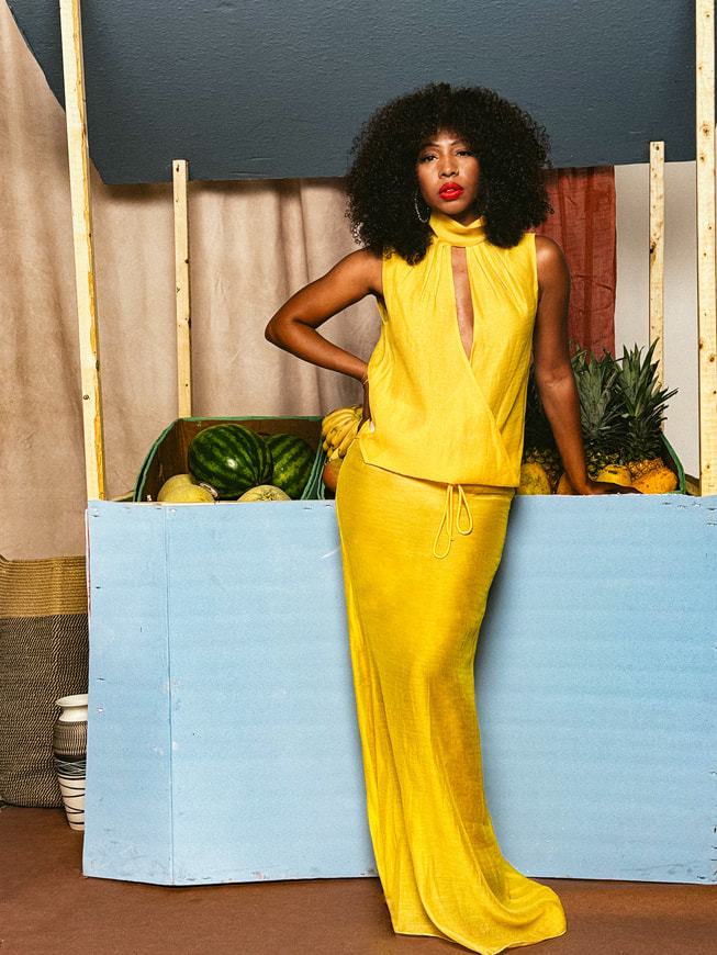 A portrait of Sammy B designer Samantha Black, wearing vibrant yellow against a backdrop made to look like a fruit stand. Shot on iPhone 16 Pro Max by Karston Tannis.