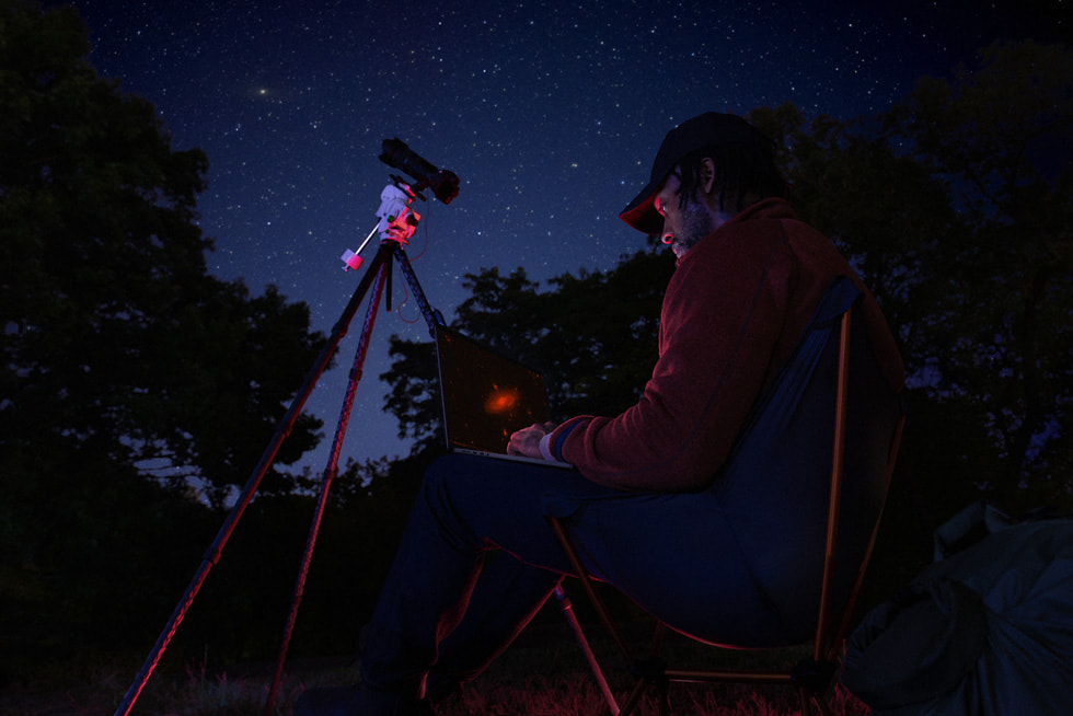 A MacBook Pro user sits outside with a telescope under a starry night’s sky.