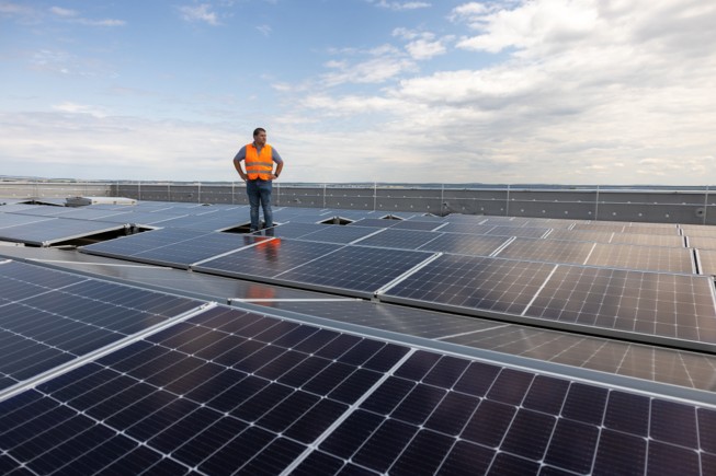 An array of solar panels is situated near a community and forested mountains.