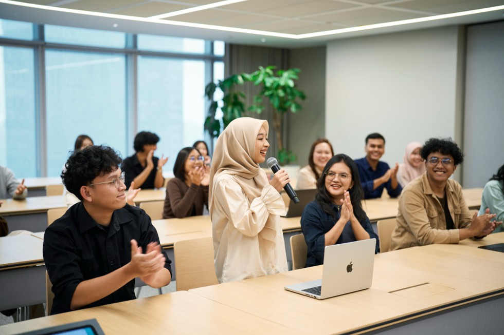 Kelompok pelajar yang beragam berkolaborasi dan tertawa dalam ruangan kelas modern di Apple Developer Institute for Professionals di Indonesia. Sebuah MacBook berada di atas meja, di depan peserta yang memegang mikrofon.