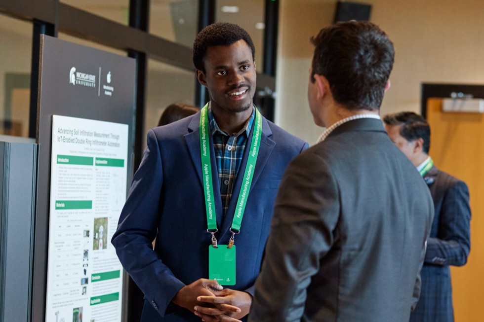 Dos participantes conversan de pie frente a un póster en el Spring Forum de la Apple Manufacturing Academy.