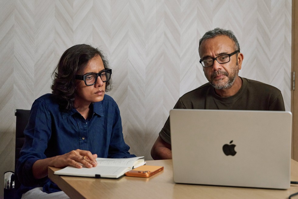 Dibakar Banerjee and Dhritisree Sarkar sit at a table, looking at a MacBook Pro. iPhone 17 Pro Max sits on the table beside them.