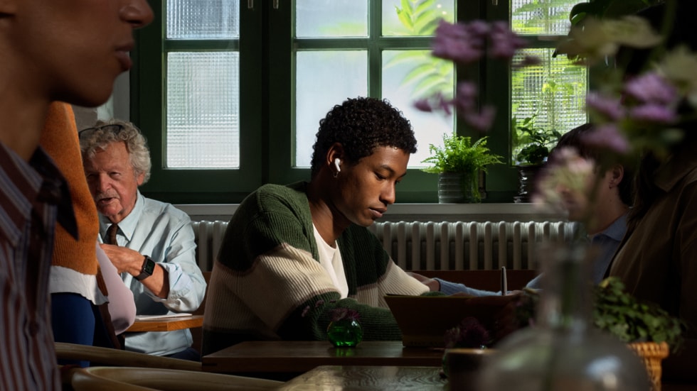 An AirPods 5 user is pictured working at a restaurant surrounded by other people.