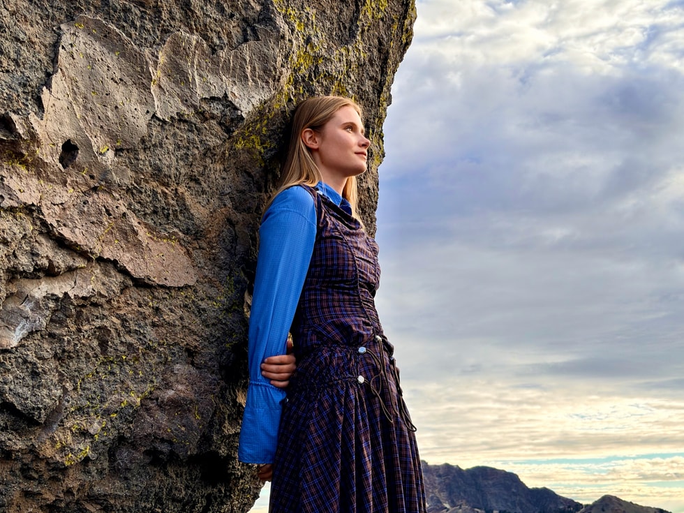 A person in a dark plaid dress stands outdoors against a tall rock formation. 