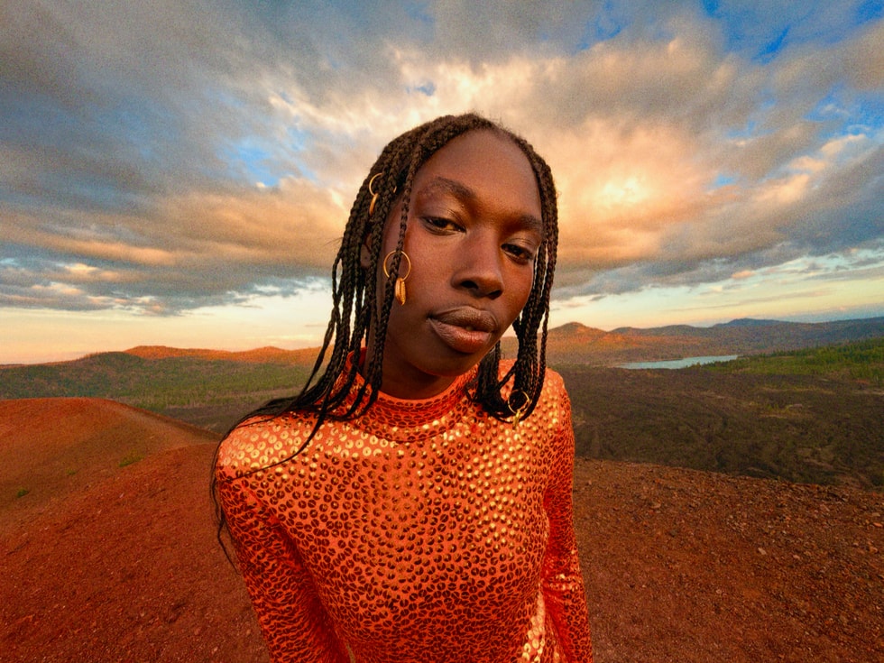 A person wearing orange poses in a semi-close-up against a cloudy sky and a wide sweeping landscape.