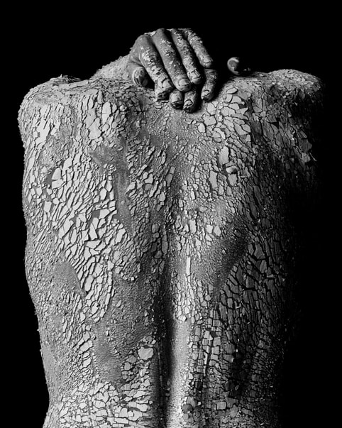 A person’s back covered in clay with their hands resting on the back of their neck.