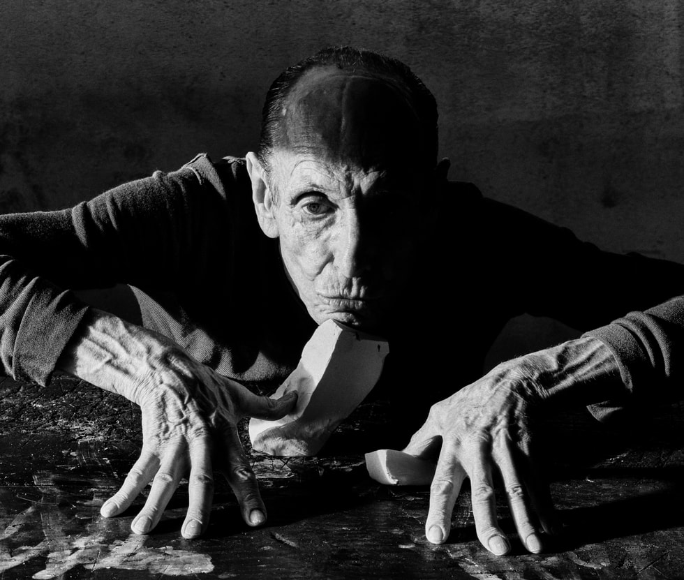 A portrait shows a person with their hands perched on a tabletop among pieces of clay.