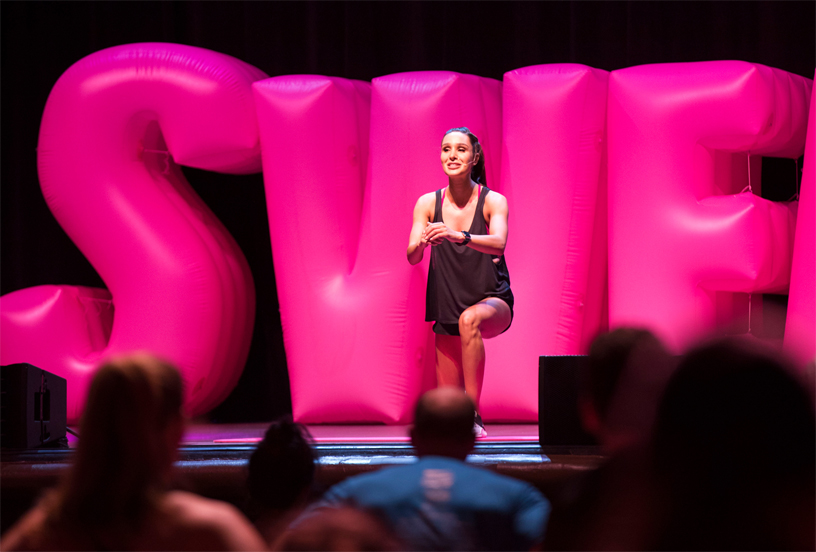 Kayla Itsines doing a fitness routine in front of inflated balloon letters on stage.