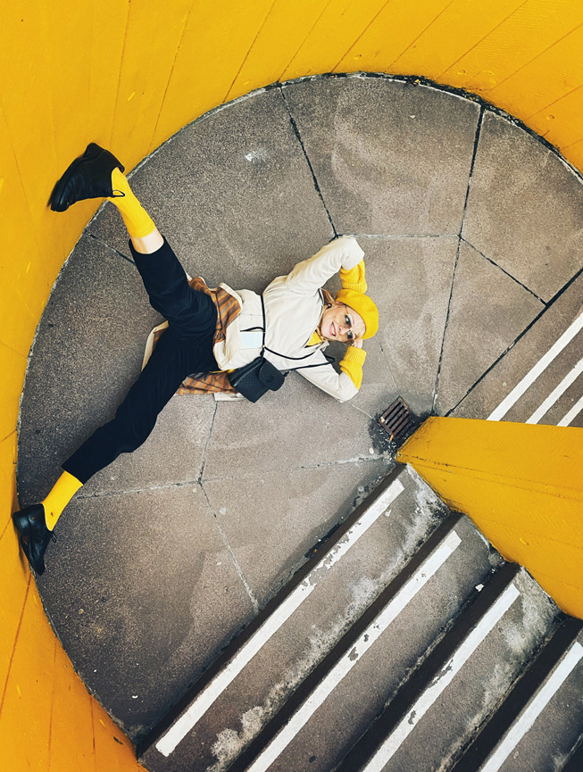 A person poses on the landing of a yellow-walled stairwell.