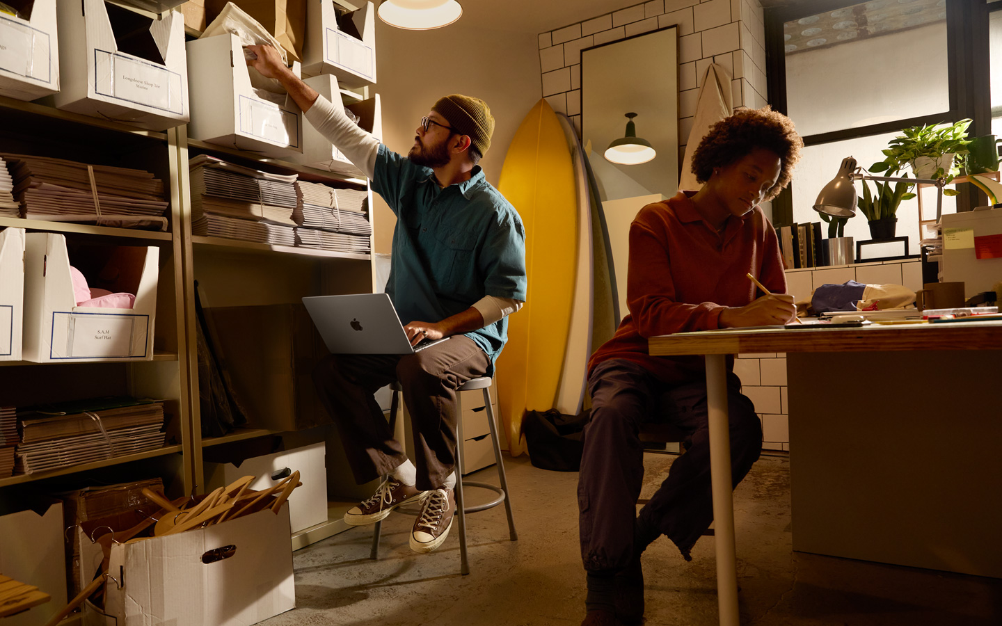 Egy ember íróasztalnál ül, és ceruzával jegyzetfüzetbe ír, egy másik ember polc mellett ül, ölében MacBook, jobb kezével egy aktáért nyúl.