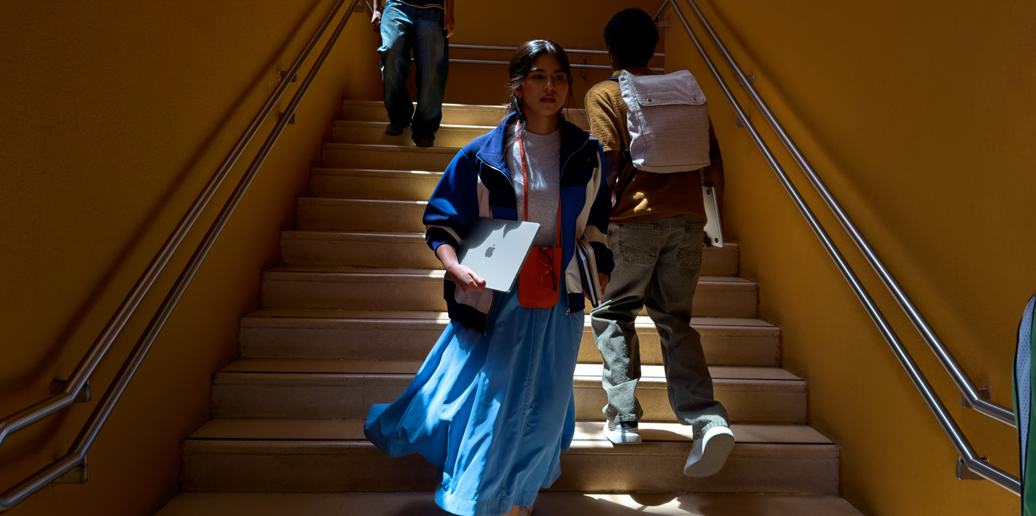 A student walking down the stairs. She is holding a MacBook and books.