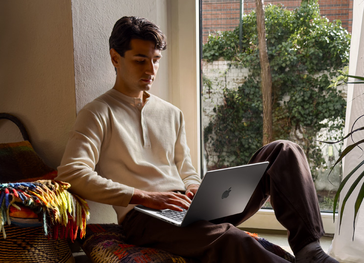 En person som sitter på golvet och använder en MacBook bredvid ett fönster.