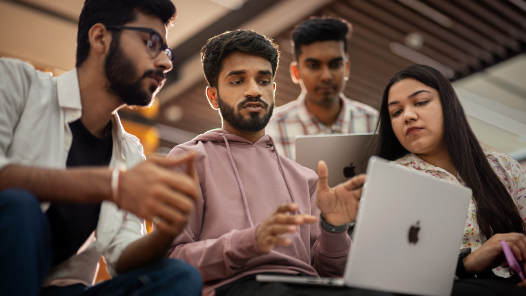 A group of four young students gathered around a MacBook engaged in collaborative discussion.