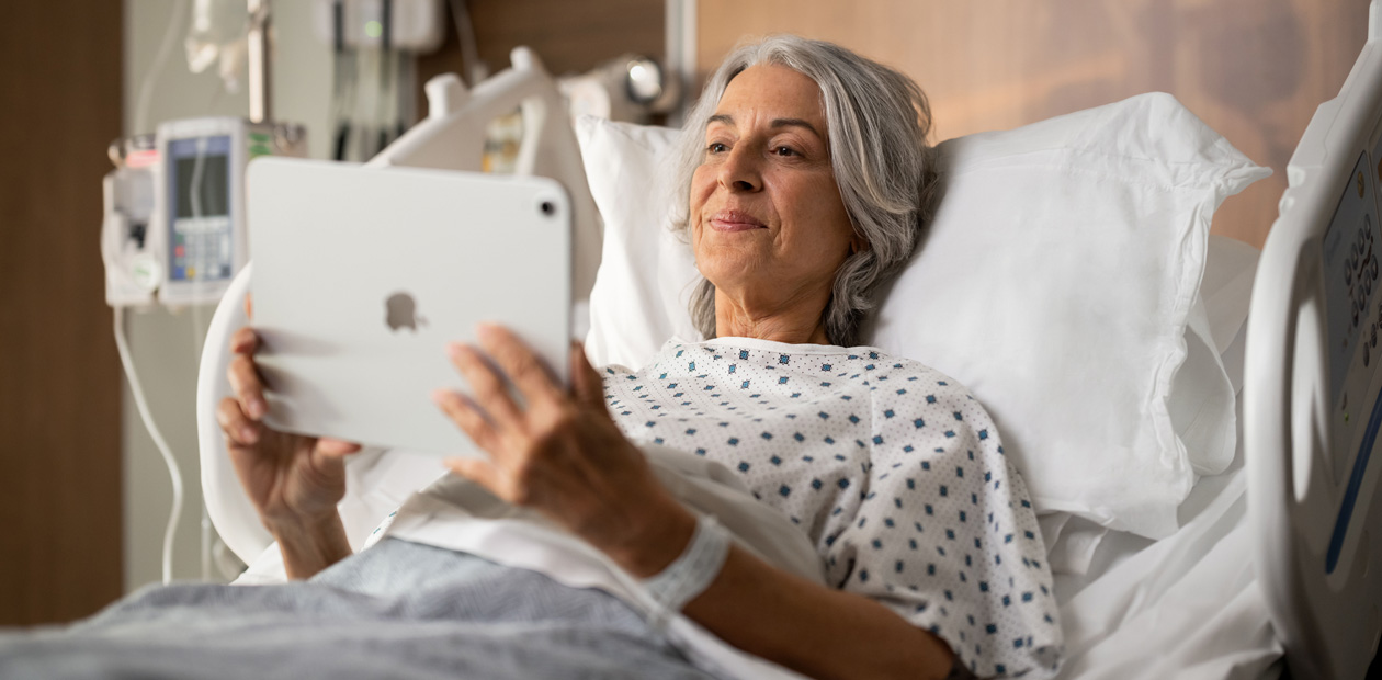 A female patient lies in a hospital bed while looking at her iPad.