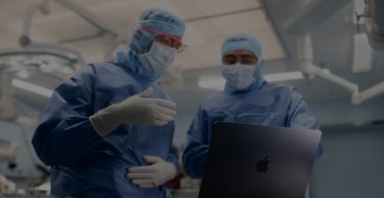 Dois cirurgiões de uniforme olham para um MacBook Pro na sala de cirurgia.