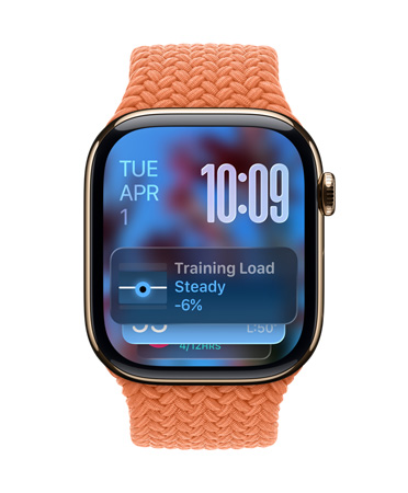 Apple Watch in Rose Gold met een gevlochten bandje in Tangerine, de wijzerplaat toont de datum linksboven, rechtsboven staat de tijd met gestapelde tijdsnotering, de Trainingsbelasting-widget staat onderaan, en alles heeft het doorschijnende Liquid Glass-design