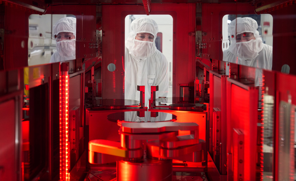 An employee with a safety suit on peers into the window of a silicon manufacturing machine